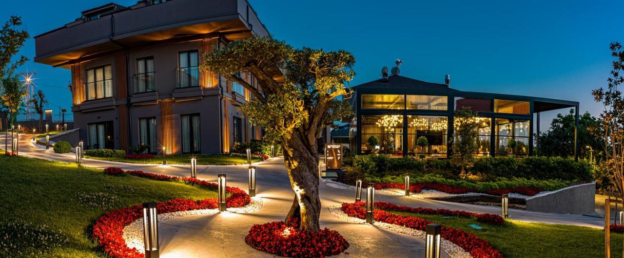 Menalo Hotel Premium Istanbul Airport Екстериор снимка The photo depicts an elegant outdoor setting at dusk. In the foreground, there's a beautifully lit olive tree, adding a natural focal point. Surrounding it are well-maintained flower beds with vibrant red flowers, arranged along a path that winds thr
