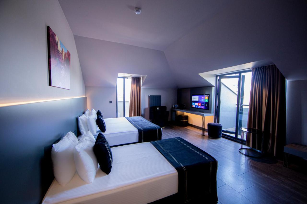 Menalo Hotel Premium Istanbul Airport Екстериор снимка The photo shows a modern hotel room with a sleek design. There are two beds with white linens and dark blue accents. A large flat-screen TV is mounted on the wall, and there is a small seating area with a chair and a table. The room features large wi